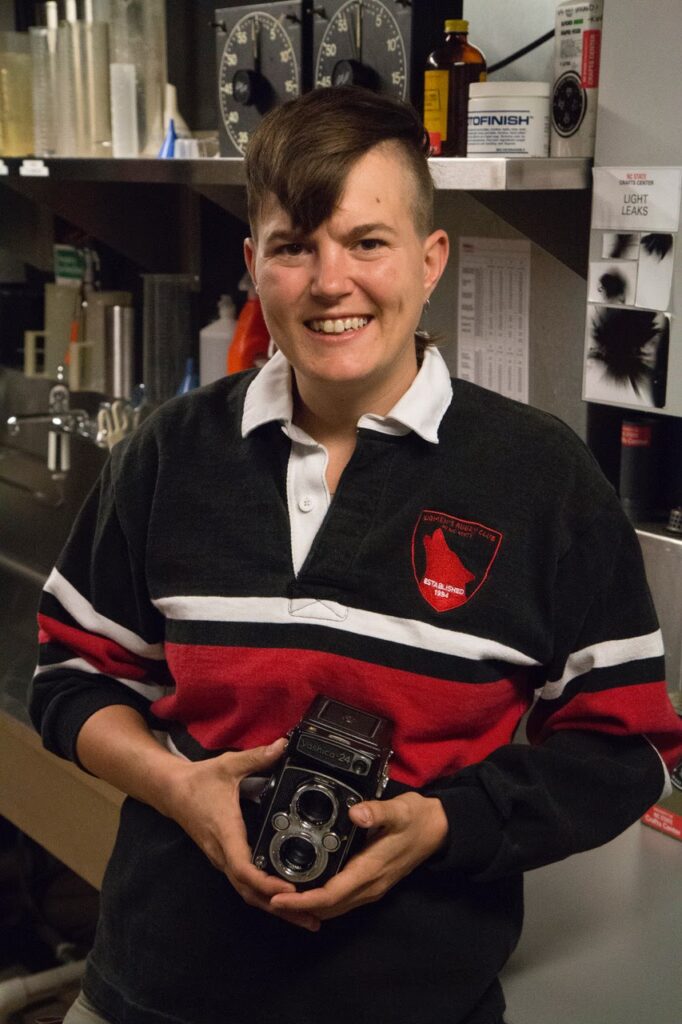 Katy smiles at the camera and holds an old fashioned camera in her hands. She is a white cis gender lesbian woman with masculine leaning, androgenous style/tastes. She has short, straight brown hair, light brown eyes, and dimple crested cheeks.