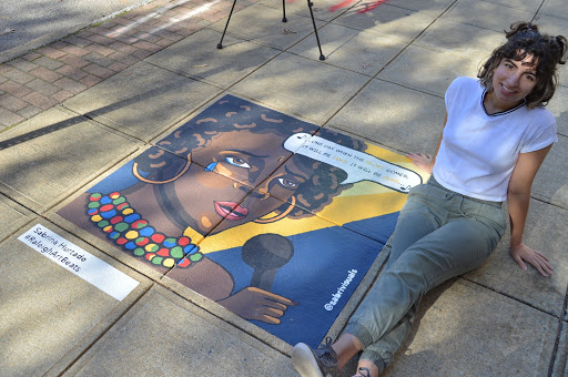 The artist, Sabrina sits next to her sidewalk mural: an image of a Black woman with hoop earrings shedding a tear painted with bold colors. There is dialog bubble that says "One day when the Glory comes, it will be ours...it will be ours"