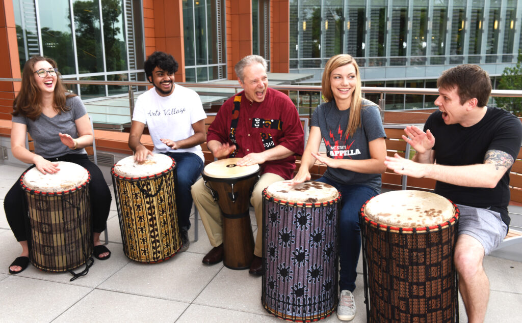 Rich Holly, executive director of the arts at NC State, sits in the center of a drum circle gathering on a balcony at Talley Student Union. He is joined by four NC State students.