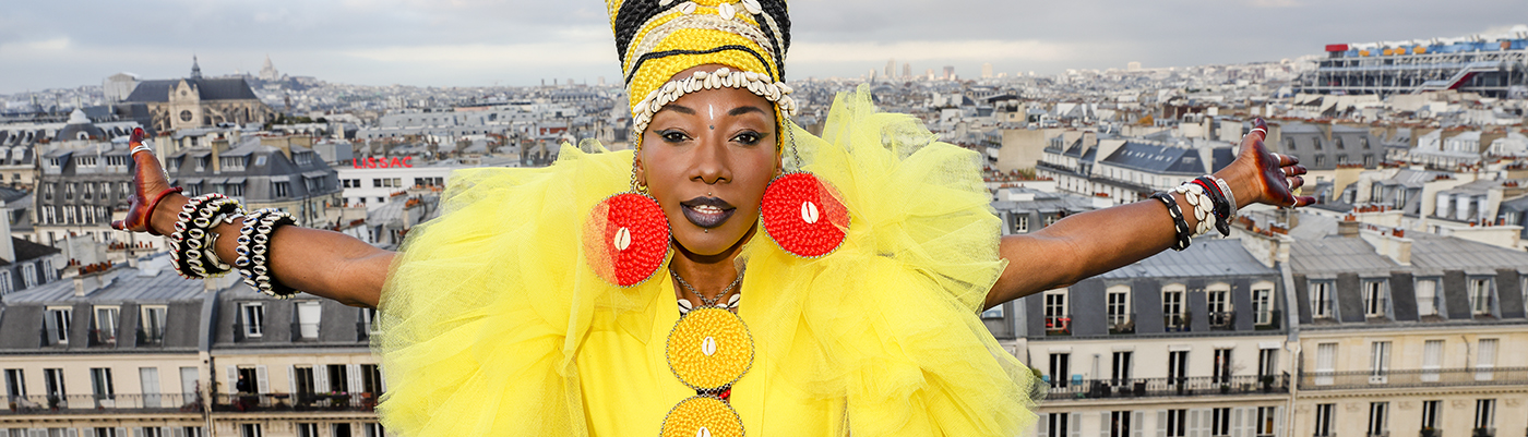 Malian guitarist and singer Fatoumata Diawara. She is wearing a bright yellow dress and multiple jewelry items made from shells. She is standing in a high spot outdoors with a city behind her.
