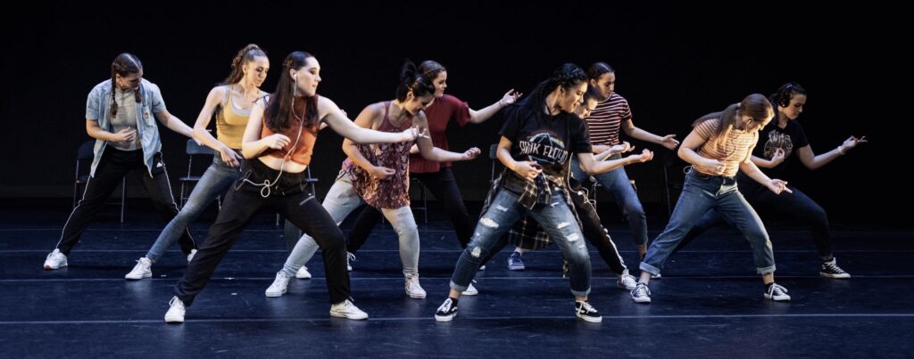 Nine members of the State Dance Company are on a stage with a dark background playing air guitar. 