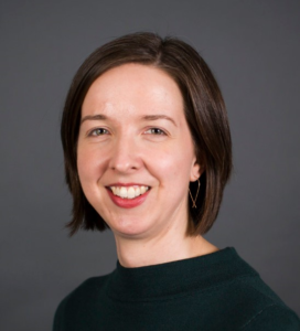 A studio photograph of Mary, a light-skinned woman with chin-length brown hair and green eyes wearing a dark green shirt and gold wire earrings and smiling at the camera in front of a gray background.