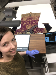 Mary stands in front of a brown desk on which a document viewer sits. The viewer is pointed at a purple and gold Chinese garment on the table behind the desk, and an image of the garment is shown on the laptop screen to the right of the desk. She is wearing nitrile gloves and is looking up at the camera and smiling. no photo credit necessary