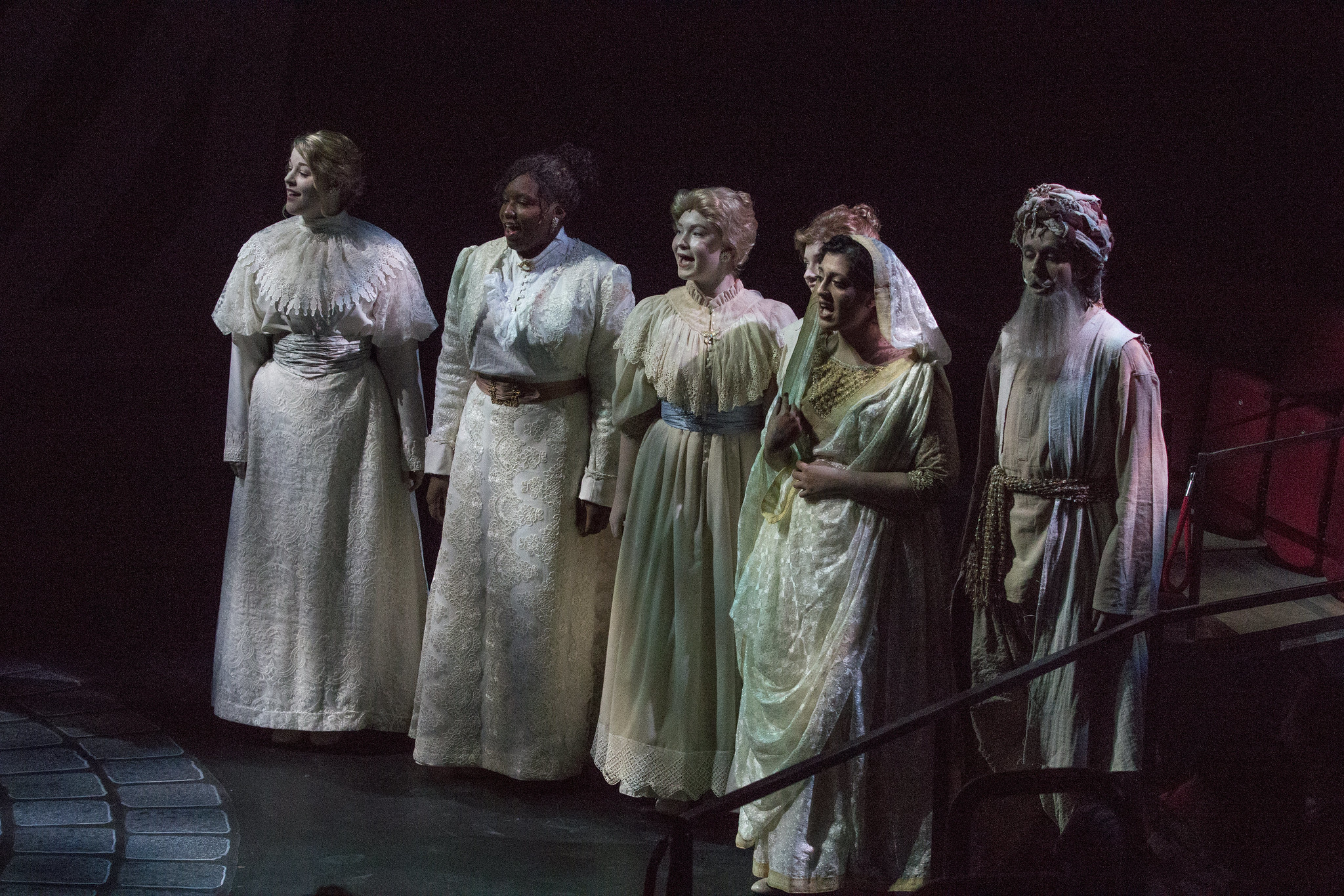 Five people are singing on a stage with shadowy light. They wear white Victorian era dresses.